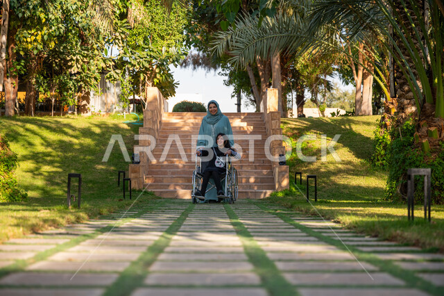 مساعدة مرضى الإعاقة الحركية على التنقل بسهولة, الاستمتاع بقضاء الوقت الممتع بالهواء الطلق, العطف والحب بين أفراد الأسرة, حديقة ومنتزه ترفيهي, امرأة عربيه خليجيه سعودية محجبة ترتدي عباءة زرقاء برفقة ابنتها من ذوي الهمم في إحدى الحدائق العامة
