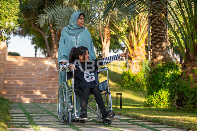 Enjoying a good time outdoors, kindness and love between family members, a garden and an amusement park, a veiled Saudi Arabian Gulf woman wearing a blue abaya with her daughter with special needs in a public park, helping patients with motor disabilities move easily