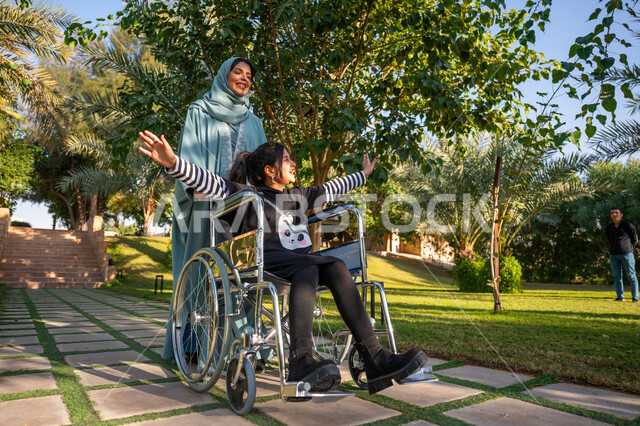 الاستمتاع بقضاء الوقت الممتع بالهواء الطلق, العطف والحب بين أفراد الأسرة, حديقة ومنتزه ترفيهي, امرأة عربيه خليجيه سعودية محجبة ترتدي عباءة زرقاء برفقة ابنتها من ذوي الهمم في إحدى الحدائق العامة, مساعدة مرضى الإعاقة الحركية على التنقل بسهولة