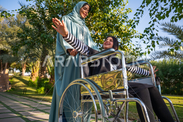 مساعدة مرضى الإعاقة الحركية على التنقل بسهولة, الاستمتاع بقضاء الوقت الممتع بالهواء الطلق, العطف والحب بين أفراد الأسرة, حديقة ومنتزه ترفيهي, امرأة عربيه خليجيه سعودية محجبة ترتدي عباءة زرقاء برفقة ابنتها من ذوي الهمم في إحدى الحدائق العامة