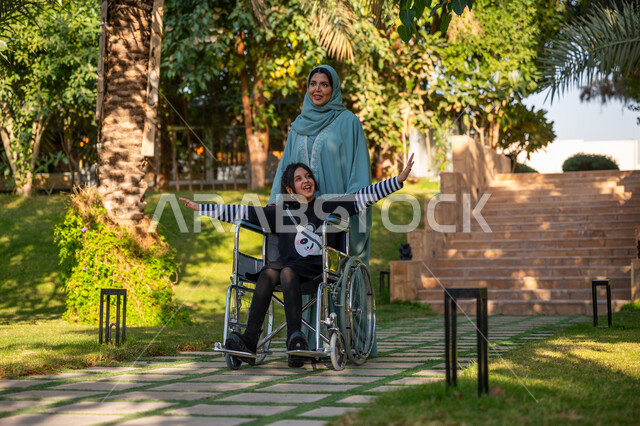 الاستمتاع بقضاء الوقت الممتع بالهواء الطلق, العطف والحب بين أفراد الأسرة, حديقة ومنتزه ترفيهي, امرأة عربيه خليجيه سعودية محجبة ترتدي عباءة زرقاء برفقة ابنتها من ذوي الهمم في إحدى الحدائق العامة, مساعدة مرضى الإعاقة الحركية على التنقل بسهولة