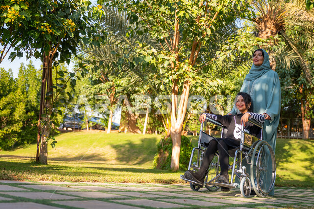 مساعدة مرضى الإعاقة الحركية على التنقل بسهولة, الاستمتاع بقضاء الوقت الممتع بالهواء الطلق, العطف والحب بين أفراد الأسرة, حديقة ومنتزه ترفيهي, امرأة عربيه خليجيه سعودية محجبة ترتدي عباءة زرقاء برفقة ابنتها من ذوي الهمم في إحدى الحدائق العامة