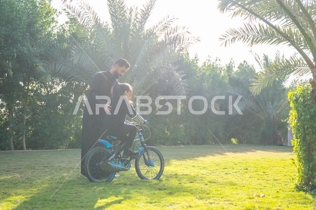 مفهوم مساندة الأب لأبنائه, حديقة ومنتزه ترفيهي عائلي, الاستمتاع بممارسة الهواية المفضلة بالهواء الطلق, رجل عربي خليجي سعودي يرتدي الثوب التقليدي يقوم بمساعدة ابنته بقيادة الدراجة الهوائية, قضاء الوقت الممتع مع العائلة