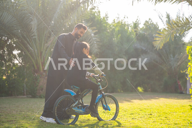 قضاء الوقت الممتع مع العائلة, مفهوم مساندة الأب لأبنائه, حديقة ومنتزه ترفيهي عائلي, الاستمتاع بممارسة الهواية المفضلة بالهواء الطلق, رجل عربي خليجي سعودي يرتدي الثوب التقليدي يقوم بمساعدة ابنته بقيادة الدراجة الهوائية