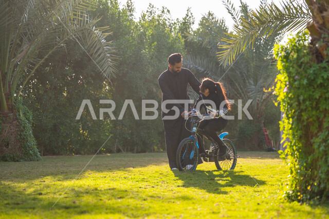 حديقة ومنتزه ترفيهي عائلي, الاستمتاع بممارسة الرياضة المفضلة, رجل عربي خليجي سعودي يرتدي الثوب التقليدي يقوم بمساعدة ابنته بقيادة الدراجة الهوائية، نشاط حركي في الهواء الطلق، قضاء الوقت الممتع مع العائلة، مفهوم مساندة الأب لأبنائه