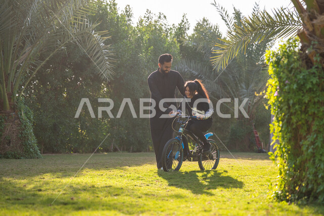 مفهوم مساندة الأب لأبنائه, حديقة ومنتزه ترفيهي عائلي, الاستمتاع بممارسة الرياضة المفضلة, رجل عربي خليجي سعودي يرتدي الثوب التقليدي يقوم بمساعدة ابنته بقيادة الدراجة الهوائية، نشاط حركي في الهواء الطلق، قضاء الوقت الممتع مع العائلة