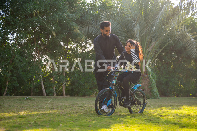 قضاء الوقت الممتع مع العائلة, مفهوم مساندة الأب لأبنائه, حديقة ومنتزه ترفيهي عائلي, الاستمتاع بممارسة الرياضة المفضلة, رجل عربي خليجي سعودي يرتدي الثوب التقليدي يقوم بمساعدة ابنته بقيادة الدراجة الهوائية، نشاط حركي في الهواء الطلق