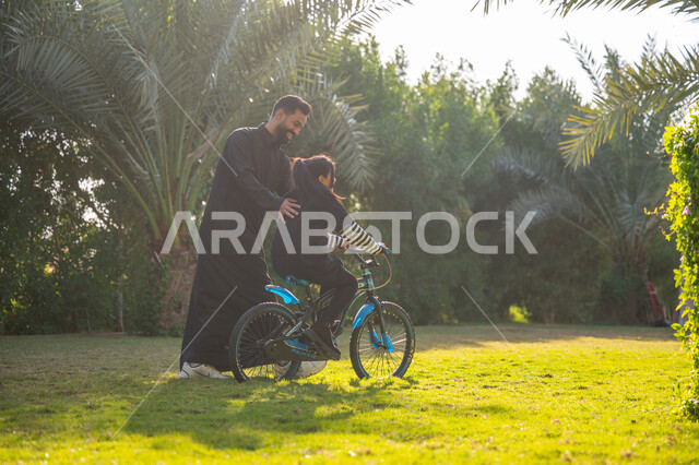 مفهوم مساندة الأب لأبنائه, حديقة ومنتزه ترفيهي عائلي, الاستمتاع بممارسة الهواية المفضلة بالهواء الطلق, رجل عربي خليجي سعودي يرتدي الثوب التقليدي يقوم بمساعدة ابنته بقيادة الدراجة الهوائية, قضاء الوقت الممتع مع العائلة