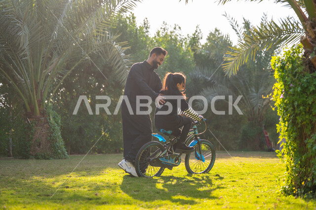 حديقة ومنتزه ترفيهي عائلي, الاستمتاع بممارسة الرياضة المفضلة, رجل عربي خليجي سعودي يرتدي الثوب التقليدي يقوم بمساعدة ابنته بقيادة الدراجة الهوائية، نشاط حركي في الهواء الطلق، قضاء الوقت الممتع مع العائلة، مفهوم مساندة الأب لأبنائه