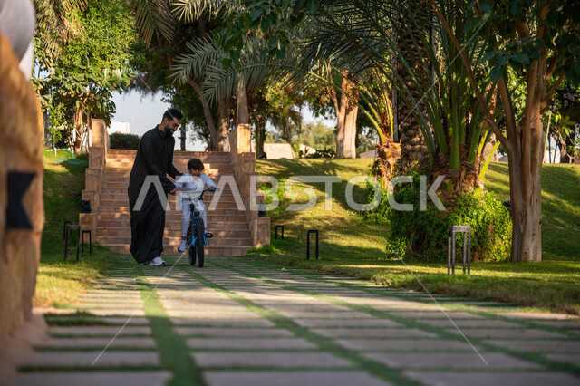 مفهوم مساندة الأب لأبنائه, حديقة ومنتزه ترفيهي عائلي, الاستمتاع بممارسة الهواية المفضلة بالهواء الطلق, رجل عربي خليجي سعودي يرتدي الثوب التقليدي يقوم بمساعدة ابنه بقيادة الدراجة الهوائية, قضاء الوقت الممتع مع العائلة