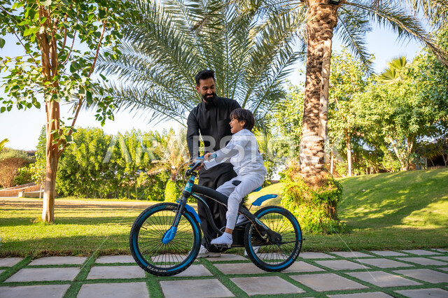 قضاء الوقت الممتع مع العائلة, مفهوم مساندة الأب لأبنائه, حديقة ومنتزه ترفيهي عائلي, الاستمتاع بممارسة الهواية المفضلة بالهواء الطلق, رجل عربي خليجي سعودي يرتدي الثوب التقليدي يقوم بمساعدة ابنه بقيادة الدراجة الهوائية