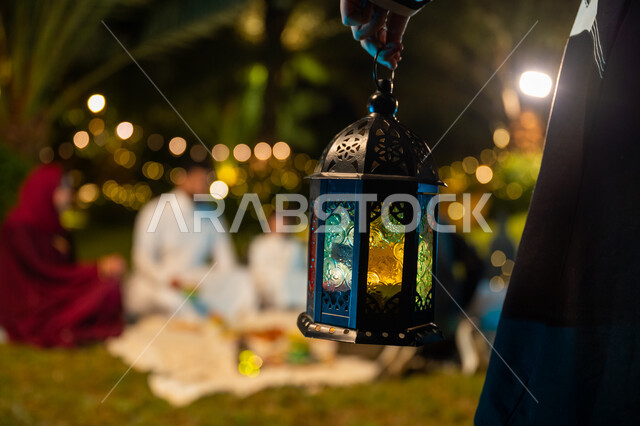 أجواء عائلية سعيدة في شهر رمضان المبارك، التسامر وتبادل الأحاديث بين الأفراد، أسرة عربية خليجية سعودية تقضي وقت ممتع في جلسة خارجية في احدى الشاليهات، صورة مقربة ليد فتاة تحمل فانوس معدني مضيء، ديكور وزينة المناسبات والأعياد الاسلامية