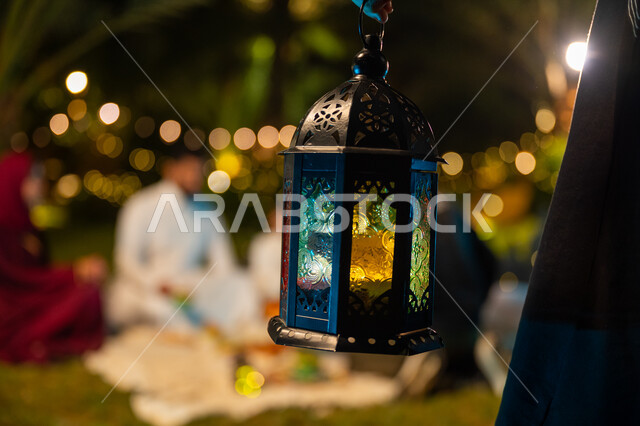 التسامر وتبادل الأحاديث بين الأفراد، أسرة عربية خليجية سعودية تقضي وقت ممتع في جلسة خارجية في احدى الشاليهات، أجواء عائلية سعيدة في شهر رمضان المبارك، صورة مقربة ليد فتاة تحمل فانوس معدني مضيء، ديكور وزينة المناسبات والأعياد الاسلامية