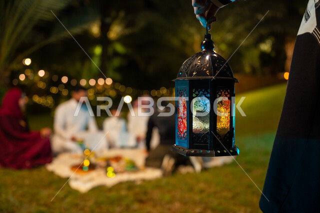 تبادل الأحاديث والتسامر بين الأفراد، أسرة عربية خليجية سعودية تقضي وقت ممتع في جلسة خارجية في احدى الشاليهات، أجواء عائلية سعيدة في شهر رمضان المبارك، صورة مقربة ليد فتاة تحمل فانوس معدني مضيء، ديكور وزينة المناسبات والأعياد الاسلامية