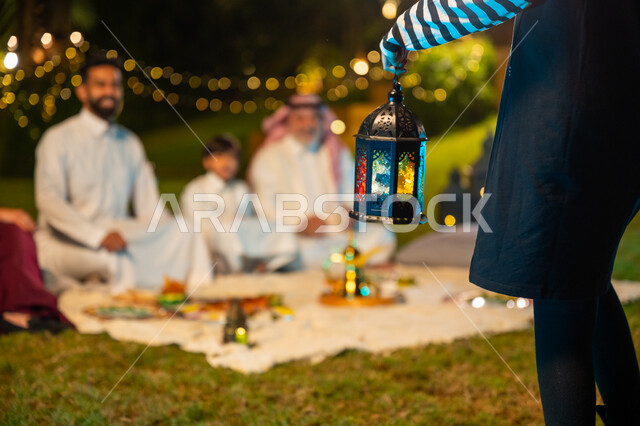 ديكور وزينة المناسبات والأعياد الاسلامية، ايماءات السعادة والسرور بالاجتماع العائلي، أجواء عائلية سعيدة في شهر رمضان المبارك، أسرة عربية خليجية سعودية تقضي وقت ممتع في جلسة خارجية في احدى الشاليهات، صورة مقربة ليد فتاة تحمل فانوس معدني مضيء