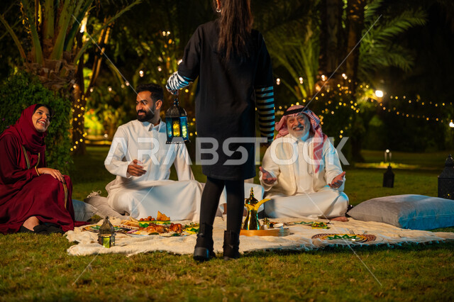 أجواء عائلية سعيدة في شهر رمضان المبارك، زينة وديكور المناسبات والأعياد الاسلامية، ايماءات السعادة والسرور بالاجتماع العائلي، أسرة عربية خليجية سعودية تقضي وقت ممتع في جلسة خارجية في احدى الشاليهات، صورة من الخلف لفتاة تحمل بيدها فانوس معدني مضيء