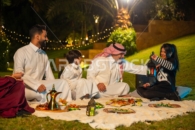 زينة وديكور المناسبات والأعياد الاسلامية، أجواء عائلية سعيدة في شهر رمضان المبارك، ايماءات السعادة والسرور بالاجتماع العائلي، أسرة عربية خليجية سعودية تقضي وقت ممتع في جلسة خارجية في احدى الشاليهات، فتاة تحمل بيدها فانوس معدني مضيء