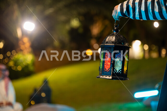 Spiritual Ramadan night atmosphere, decorations and accessories for Islamic holidays and occasions, a close-up of a Saudi Arabian Gulf girl's hand holding a decorated illuminated metal lantern, spending a pleasant time in an outdoor family session in one of the chalets, welcoming the holy month of Ramadan