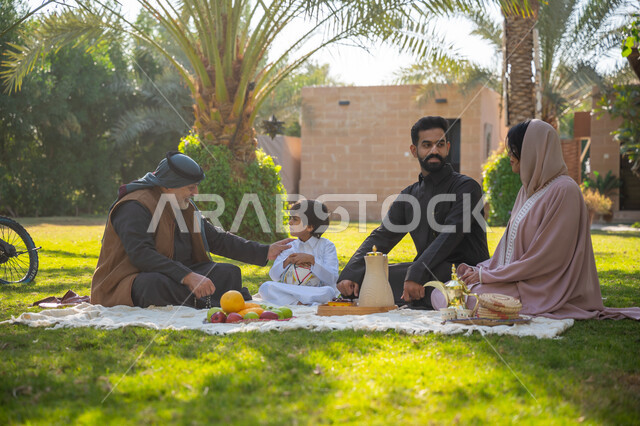 تبادل الأحاديث بإيماءات السرور، عائلة عربية خليجية سعودية يرتدون الزي التقليدي يجلسون في إحدى الحدائق العامة في النهار, قضاء الوقت الممتع مع الأسرة, حديقة ومنتزه عائلي ترفيهي, الاستمتاع بالنزهة في الهواء الطلق, تقوية وتعزيز الروابط الأسرية