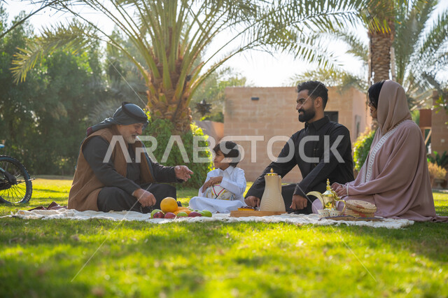 تقوية وتعزيز الروابط الأسرية، تبادل الأحاديث بإيماءات السرور، عائلة عربية خليجية سعودية يرتدون الزي التقليدي يجلسون في إحدى الحدائق العامة في النهار, قضاء الوقت الممتع مع الأسرة, حديقة ومنتزه عائلي ترفيهي, الاستمتاع بالنزهة في الهواء الطلق