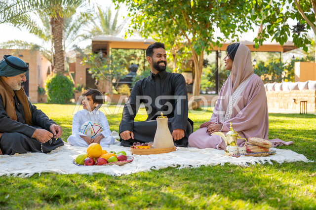 تقوية وتعزيز الروابط الأسرية، تبادل الأحاديث بإيماءات السرور، عائلة عربية خليجية سعودية يرتدون الزي التقليدي يجلسون في إحدى الحدائق العامة في النهار, قضاء الوقت الممتع مع الأسرة, حديقة ومنتزه عائلي ترفيهي, الاستمتاع بالنزهة في الهواء الطلق
