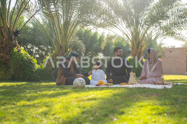 تبادل الأحاديث بإيماءات السرور، عائلة عربية خليجية سعودية يرتدون الزي التقليدي يجلسون في إحدى الحدائق العامة في النهار, قضاء الوقت الممتع مع الأسرة, حديقة ومنتزه عائلي ترفيهي, الاستمتاع بالنزهة في الهواء الطلق, تقوية وتعزيز الروابط الأسرية