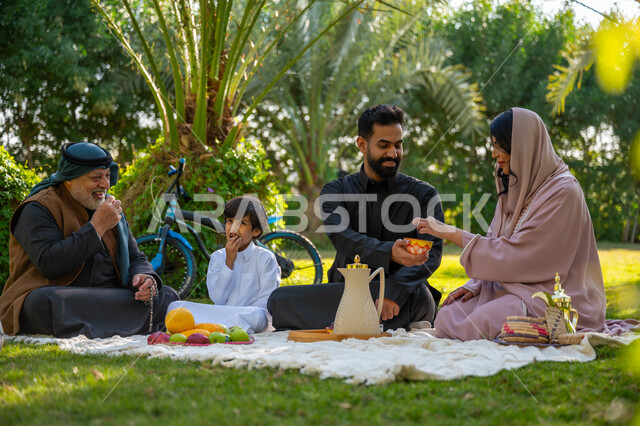 تبادل الأحاديث بإيماءات السرور، عائلة عربية خليجية سعودية يرتدون الزي التقليدي يجلسون في إحدى الحدائق العامة في النهار, قضاء الوقت الممتع مع الأسرة, حديقة ومنتزه عائلي ترفيهي, الاستمتاع بالنزهة في الهواء الطلق, تقوية وتعزيز الروابط الأسرية