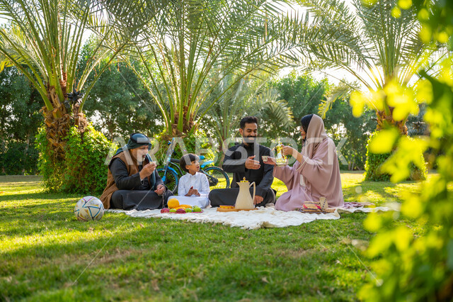حديقة ومنتزه عائلي ترفيهي, الاستمتاع بالنزهة في الهواء الطلق, تقوية وتعزيز الروابط الأسرية، تبادل الأحاديث بإيماءات السرور، عائلة عربية خليجية سعودية يرتدون الزي التقليدي يجلسون في إحدى الحدائق العامة في النهار, قضاء الوقت الممتع مع الأسرة
