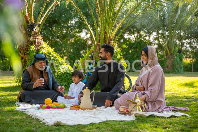 تبادل الأحاديث بإيماءات السرور, تقوية وتعزيز الروابط الأسرية، عائلة عربية خليجية سعودية يرتدون الزي التقليدي يجلسون في إحدى الحدائق العامة في النهار, قضاء الوقت الممتع مع الأسرة, حديقة ومنتزه عائلي ترفيهي, الاستمتاع بالنزهة في الهواء الطلق