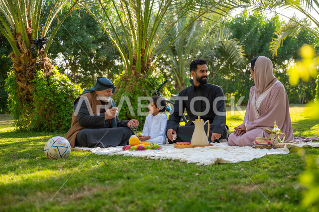 تقوية وتعزيز الروابط الأسرية، تبادل الأحاديث بإيماءات السرور، عائلة عربية خليجية سعودية يرتدون الزي التقليدي يجلسون في إحدى الحدائق العامة في النهار, قضاء الوقت الممتع مع الأسرة, حديقة ومنتزه عائلي ترفيهي, الاستمتاع بالنزهة في الهواء الطلق