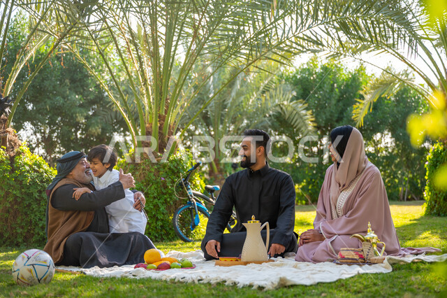 تبادل الأحاديث بإيماءات السرور, تقوية وتعزيز الروابط الأسرية، عائلة عربية خليجية سعودية يرتدون الزي التقليدي يجلسون في إحدى الحدائق العامة في النهار, قضاء الوقت الممتع مع الأسرة, حديقة ومنتزه عائلي ترفيهي, الاستمتاع بالنزهة في الهواء الطلق