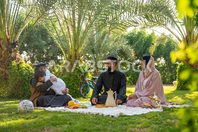 تقوية وتعزيز الروابط الأسرية، تبادل الأحاديث بإيماءات السرور، عائلة عربية خليجية سعودية يرتدون الزي التقليدي يجلسون في إحدى الحدائق العامة في النهار, قضاء الوقت الممتع مع الأسرة, حديقة ومنتزه عائلي ترفيهي, الاستمتاع بالنزهة في الهواء الطلق