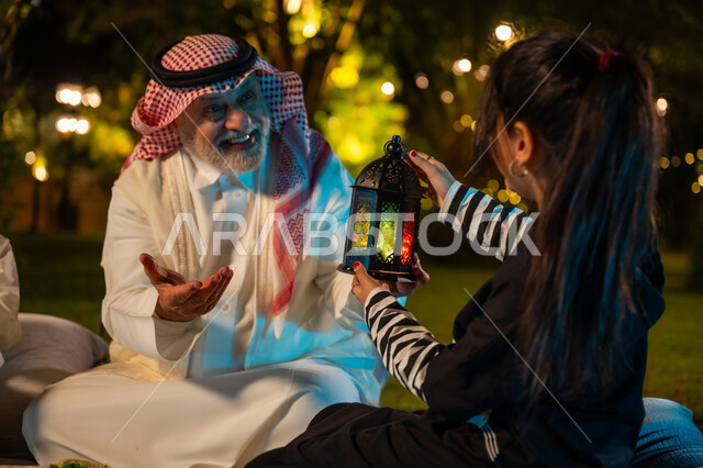 Gestures of happiness and pleasure, spending a pleasant time in one of the gardens of the luxurious chalets in Saudi Arabia, a spiritual Ramadan night atmosphere, decorations and accessories for Islamic holidays and occasions, a Saudi Arabian Gulf girl holding a lit metal lantern in her hand, sitting with her grandfather in the garden, welcoming the holy month of Ramadan