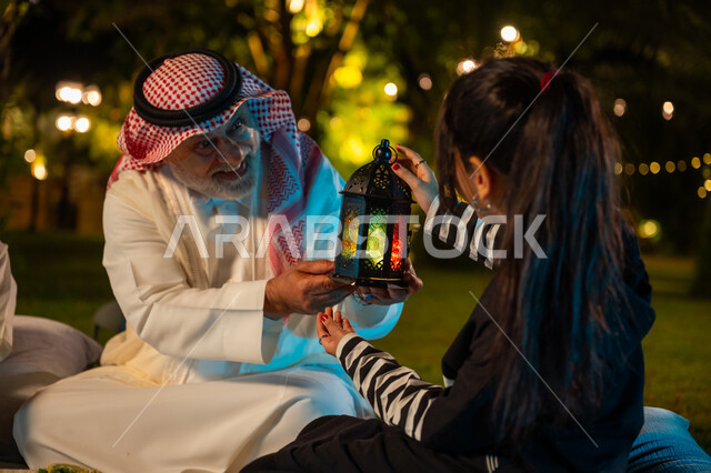 قضاء وقت ممتع في إحدى حدائق الشاليهات الفاخرة بالسعودية، أجواء ليلية رمضانية روحانية، زينة واكسسوار الأعياد والمناسبات الاسلامية، فتاة عربية خليجية سعودية تحمل بيدها فانوس معدني مضيء تجلس مع جدها في الحديقة، استقبال شهر رمضان المبارك، ايماءات السعادة والسرور