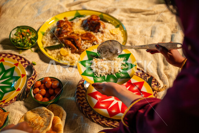 طبق من أرز المندي والدجاج المشوي اللذيذ على مائدة الإفطار، سفرة طعام بأصناف متنوعة، صورة مقربة ليدين إمرأة عربية خليجية سعودية تقوم بسكب الأطعمة الشهية، أجواء عائلية سعيدة في شهر رمضان المبارك