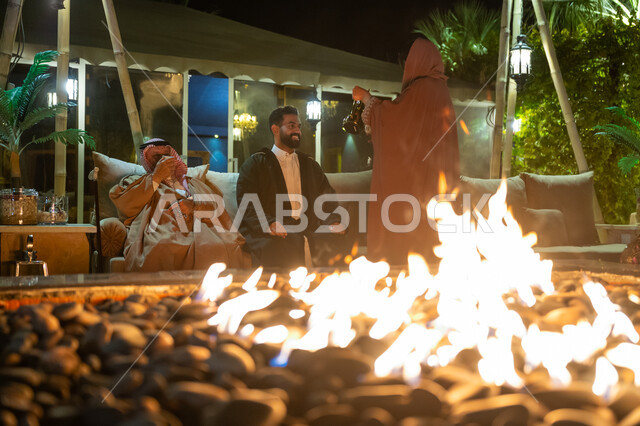 تبادل الحديث والتسامر بين الأفراد في كشتة بر في الأجواء الشتوية، حسن الضيافة والاستقبال، أسرة عربية خليجية سعودية تقضي وقت ممتع في جلسة خارجية في الشاليه، سيدة تقوم بتقديم القهوة العربية للضيوف، التدفئة على الحطب في الجو الشتوي في الليل