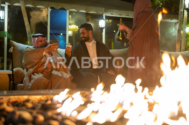 Hospitality and reception, exchanging conversation and chatting between individuals on a desert outing in the winter, a Saudi Arabian Gulf family spending a pleasant time in an outdoor session in the chalet, a woman serving Arabic coffee to guests, heating with wood in the winter at night