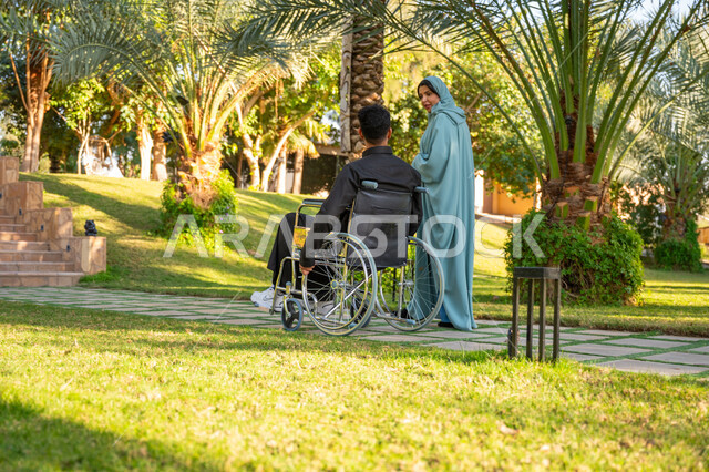 شعور ذوي الهمم بالراحة والطمأنينة في بيئتهم, مساعدة مرضى الإعاقة الحركية على التنقل بسهولة, الاستمتاع بقضاء الوقت الممتع بالهواء الطلق, صورة من الخلف لامرأة عربية خليجية سعودية محجبة ترتدي عباءة زرقاء برفقة زوجها المقعد يتنزهان في الحديقة العامة