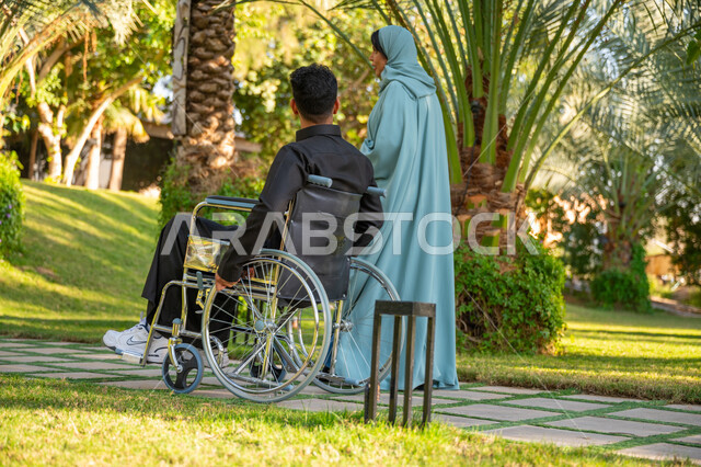 Affection and love between family members, enjoying a good time outdoors, a back view of a veiled Saudi Arabian Gulf woman wearing a blue abaya with her wheelchair husband walking in a public park, helping people with motor disabilities move around easily