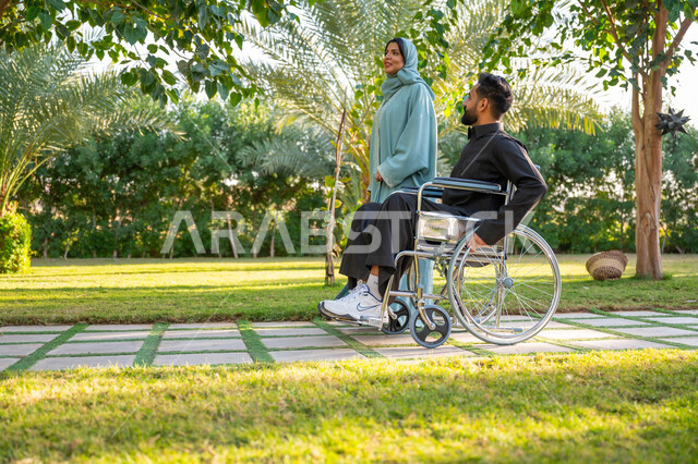 الاستمتاع بقضاء الوقت الممتع بالهواء الطلق, العطف والحب بين أفراد الأسرة, امرأة عربية خليجية سعودية محجبة ترتدي عباءة زرقاء برفقة زوجها المقعد يتنزهان في الحديقة العامة, شعور ذوي الهمم بالراحة والطمأنينة في بيئتهم, مساعدة مرضى الإعاقة الحركية على التنقل بسهولة