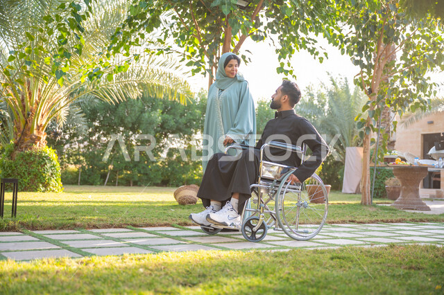 شعور ذوي الهمم بالراحة والطمأنينة في بيئتهم, مساعدة مرضى الإعاقة الحركية على التنقل بسهولة, الاستمتاع بقضاء الوقت الممتع بالهواء الطلق, العطف والحب بين أفراد الأسرة, امرأة عربية خليجية سعودية محجبة ترتدي عباءة زرقاء برفقة زوجها المقعد يتنزهان في الحديقة العامة