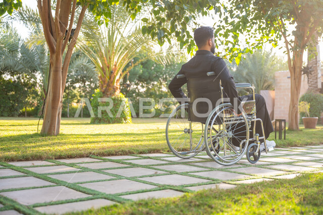 نمط حياة ذوي الهمم في المملكة, الاستمتاع بالهواء الطلق, صورة من الخلف لشاب مقعد عربي خليجي سعودي يرتدي الثوب التقليدي يجلس على كرسي متحرك ويتنزه في إحدى الحدائق العامة, قضاء الوقت الممتع في المنتزه