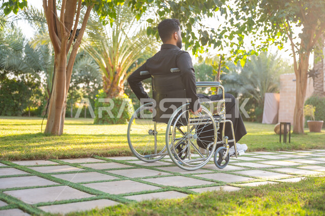 الاستمتاع بالهواء الطلق, نمط حياة ذوي الهمم في المملكة, صورة من الخلف لشاب مقعد عربي خليجي سعودي يرتدي الثوب التقليدي يجلس على كرسي متحرك ويتنزه في إحدى الحدائق العامة, قضاء الوقت الممتع في المنتزه
