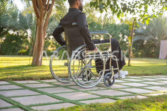 قضاء الوقت الممتع في المنتزه, الاستمتاع بالهواء الطلق, نمط حياة ذوي الهمم في المملكة, صورة من الخلف لشاب مقعد عربي خليجي سعودي يرتدي الثوب التقليدي يجلس على كرسي متحرك ويتنزه في إحدى الحدائق العامة