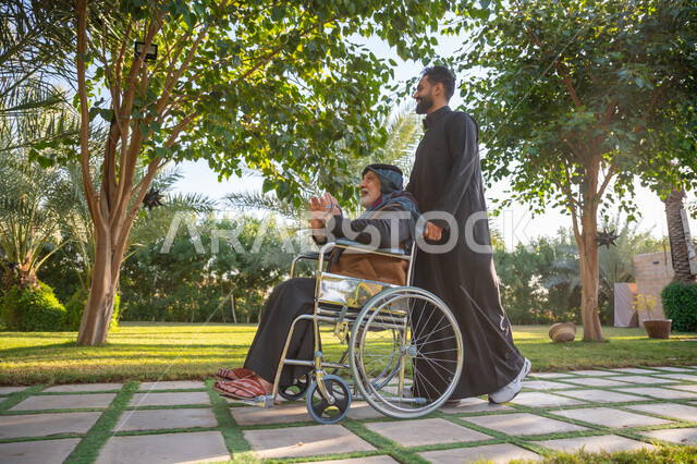 Helping people with motor disabilities move easily, Enjoying a good time outdoors, The concept of kindness, love and filial piety, A garden and amusement park, A Saudi Arabian Gulf man wearing traditional clothing with his father with special needs in a public park