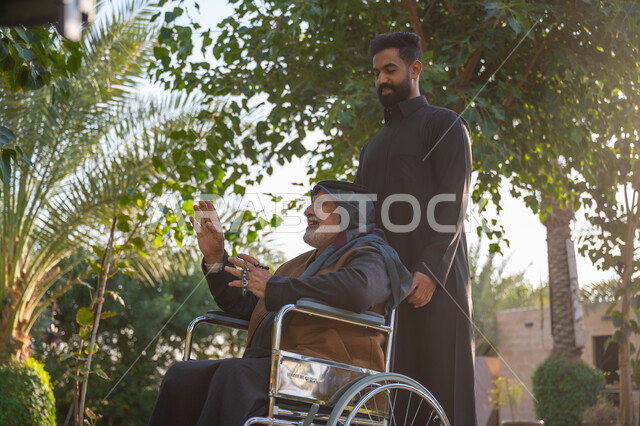 مساعدة مرضى الإعاقة الحركية على التنقل بسهولة, الاستمتاع بقضاء الوقت الممتع بالهواء الطلق, مفهوم العطف والحب وبر الوالدين, حديقة ومنتزه ترفيهي, رجل عربي خليجي سعودي يرتدي الثوب التقليدي برفقة والده من ذوي الهمم في إحدى الحدائق العامة
