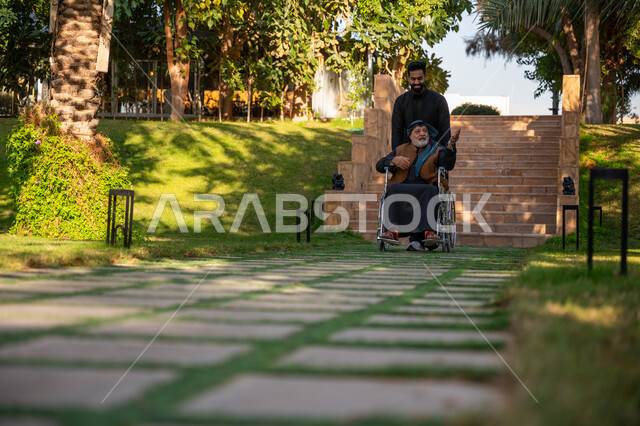 Helping people with motor disabilities move easily, Enjoying a good time outdoors, The concept of kindness, love and filial piety, A garden and amusement park, A Saudi Arabian Gulf man wearing traditional clothing with his father with special needs in a public park