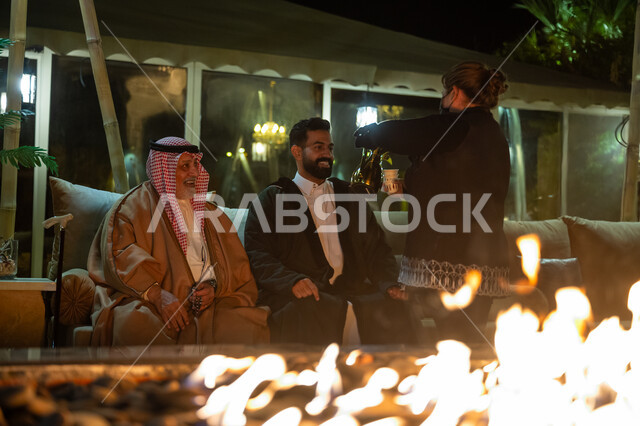 حسن الضيافة والاستقبال، العادات والتقاليد الأصيلة، التسامر وتبادل الأحاديث بين أفراد العائلة، عاملة أجنبية تقوم بتقديم القهوة العربية للضيوف، رجلان عربيان خليجيان سعوديان يجلسان بالقرب من موقد النار في الأجواء الباردة في الليل، قضاء وقت ممتع مع الأصدقاء