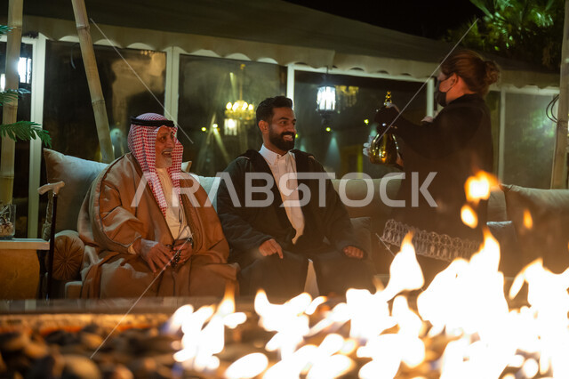 العادات والتقاليد الأصيلة، حسن الضيافة والاستقبال، التسامر وتبادل الأحاديث بين أفراد العائلة، عاملة أجنبية تقوم بتقديم القهوة العربية للضيوف، رجلان عربيان خليجيان سعوديان يجلسان بالقرب من موقد النار في الأجواء الباردة في الليل، قضاء وقت ممتع مع الأصدقاء