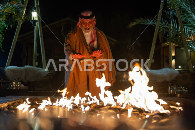 قضاء وقت ممتع في الإجازة، جلسة وكشتة بر في منطقة طبيعية بالمملكة العربية السعودية، إشعال موقد الحطب للتدفئة، الأجواء الشتوية الباردة، رجل عربي خليجي سعودي يرتدي الزي التقليدي والشماغ والفروة في الجو البارد يقف أمام شعلة النار